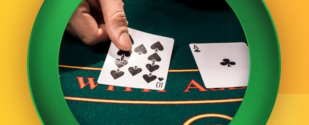 A hand holding a 10 of spades on a green blackjack table, with an ace of clubs lying face up beside it. The cards are placed on a felt surface, framed by green circular frame on a bright yellow background.