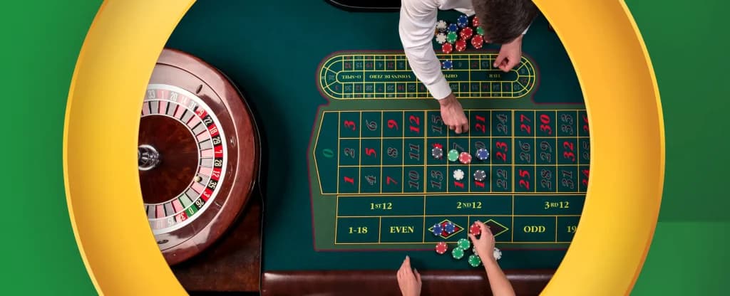 An overhead view of a roulette table showing a dealer and a player placing bets with colourful chips on red and black numbered squares. The roulette wheel, made of polished wood with red, black, and green pockets, sits on the left side. The scene is framed by a yellow circular frame on a bright green background.