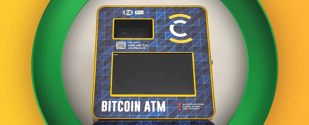 A close up view of a blue and yellow Bitcoin ATM machine with a digital screen and camera panel. The machine displays the text “BITCOIN ATM” at the bottom, along with a warning label that reads “No keys in store. Police alarm. GPS tracker.” The ATM is framed within a green circular frame on a bright yellow background.